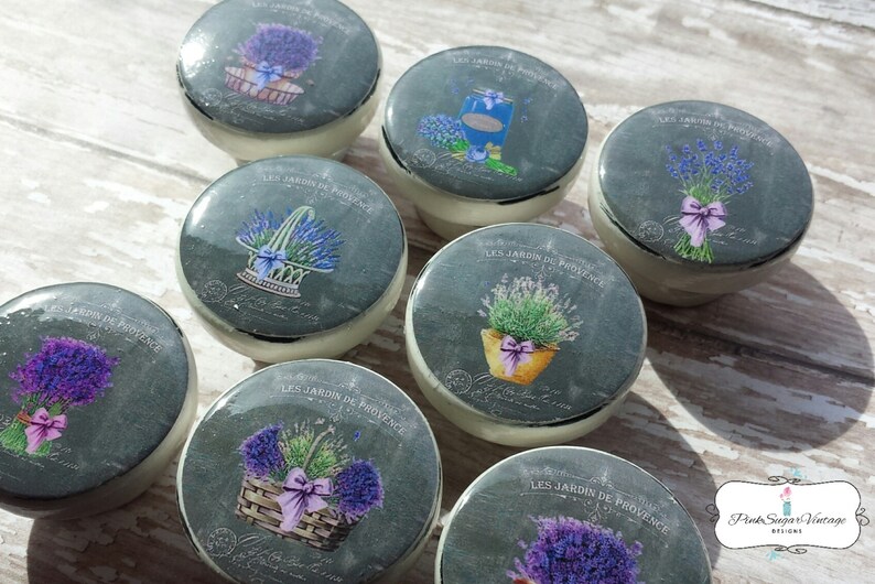 K&ouml;nnte beinhalten: Acht Keramikkn&ouml;pfe mit grauem Hintergrund und einem Lavendel-Design. Die Kn&ouml;pfe zeigen verschiedene Lavendelmotive, darunter Str&auml;u&szlig;e, K&ouml;rbe und Topfpflanzen. Der Text "LES JARDIN DE PROVENCE" ist auf jedem Knopf aufgedruckt.