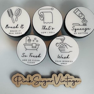 May include: Five round white ceramic bathroom themed coasters with black outlines and text. The coasters feature illustrations of a toilet paper roll, a toothbrush and toothpaste, a bathtub, a hand washing under a faucet, and a hairbrush and comb. The text on the coasters reads "That's How I Roll", "Squeeze My Bottom", "So Fresh And So Clean, Clean", "Wash Your Worries Away", and "Brush It Real Good". The coasters are arranged in a circle around a wooden sign that reads "Pink Sugar Vintage".