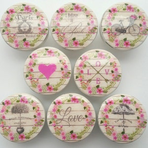 以下が含まれることがあります： 花柄とピンクのアクセントが付いた、丸い白いセラミックノブ8個セット。各ノブは、自転車、ハート、矢印に「LOVE」の文字、そして「Paris Flower Market」の文字が入った花柄など、異なるデザインが施されています。