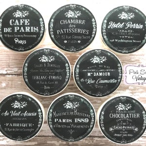 Könnte beinhalten: Neun runde schwarze und weiße Keramikknöpfe mit französisch inspirierten Designs. Die Knöpfe zeigen Text in französischer Sprache, darunter die Namen Pariser Geschäfte und Orte. Die Knöpfe sind mit floralen Motiven verziert und haben eine Vintage-Ästhetik.