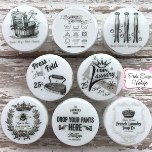 May include: Set of 8 white ceramic knobs with black and white vintage laundry themed designs. The designs include a laundry care guide, a vintage washing machine, a clothes iron, a crown with the words "Queen Bee Laundry Co.", a crown with the words "Paris, France French Laundry Soap Co.", a crown with the words "Coin Laundry", a clothes peg with the words "Keep Calm & Launder On", and a hand pointing to the words "Drop Your Pants Here".