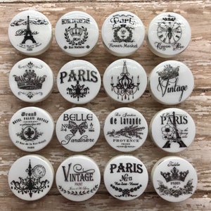 May include: Twelve white ceramic knobs with black and white Parisian themed designs. The designs include a crown, a chandelier, a Eiffel Tower, a bee, a flower market, and a vintage paint can.
