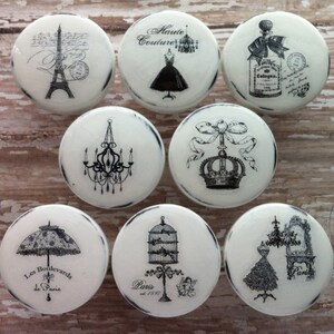 May include: Set of 8 white ceramic knobs with black and white vintage Parisian themed designs. The designs include a chandelier, a crown, a birdcage, a dress, a perfume bottle, an umbrella, and the Eiffel Tower.