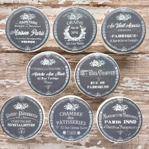 Op de afbeelding: Negen ronde grijze en witte keramische knoppen met vintage Franse tekst en bloemmotieven. De tekst omvat "Maison Paris", "Grains", "Au Vieil Acacia", "Marche Aux Fleur", "Sree Paul Charvet", "Petite Pâtisserie", "Chambre des Pâtisseries" en "Manufacture Parisienne Paris 1889".