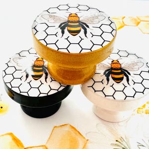 以下が含まれることがあります： 白い背景と黒いハニカム模様の丸い木製ノブが3つ。各ノブには、黄色と黒の蜂が描かれており、一番上のノブには「Bees are life」という文字が書かれています。