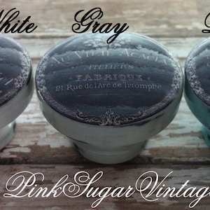 K&ouml;nnte beinhalten: Drei vintage-artige Schrankkn&ouml;pfe mit einem schwarz-wei&szlig;en Design. Die Kn&ouml;pfe sind in Wei&szlig;, Grau und Blau lackiert. Der Text auf den Kn&ouml;pfen lautet "PAPETERIE J. Ravier", "ATELIER FABRIQUE" und "DETOMME".