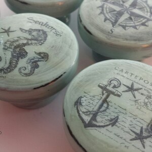 May include: Set of five decorative drawer pulls with a nautical theme. Each pull features a different design, including a compass, a seahorse, an anchor, and a postcard. The pulls are painted in a light blue color with black accents.
