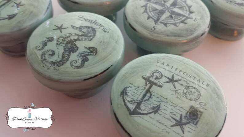 May include: Set of five decorative drawer pulls with a nautical theme. Each pull features a different design, including a compass, a seahorse, an anchor, and a postcard. The pulls are painted in a light blue color with black accents.