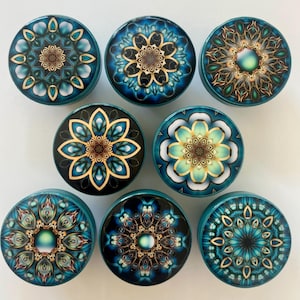 May include: A collection of nine teal and blue decorative plugs with intricate mandala designs. Each plug features a different floral pattern in shades of gold, teal, and white. The plugs are round and appear to be made of a glossy material.