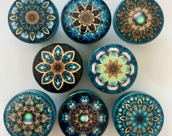 May include: A collection of nine teal and blue decorative plugs with intricate mandala designs. Each plug features a different floral pattern in shades of gold, teal, and white. The plugs are round and appear to be made of a glossy material.