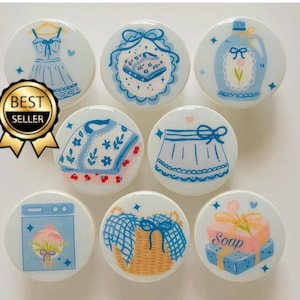 May include: Eight white ceramic phone grips with blue and pink illustrations of household items. The illustrations include a dress, a cardigan, a skirt, a bottle of soap, a laundry basket, a bar of soap, a washing machine, and a bib. The text "Soap" is visible on one of the grips.