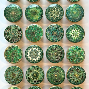 Peut inclure: Ensemble de vingt-quatre boutons de meuble ronds. Chaque bouton présente un motif floral vert détaillé avec des accents dorés. Les boutons sont disposés en grille, mettant en valeur la variété des motifs.