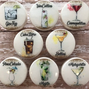 Op de afbeelding: Set van 8 ronde witte onderzetters met aquarel illustraties van verschillende cocktails. Elke onderzetter heeft de naam van de cocktail, zoals "Gin Fizz", "Tom Collins", "Cosmopolitan", "Cuba Libre", "Bellini", "Pina Colada", "Mojito" en "Margarita".