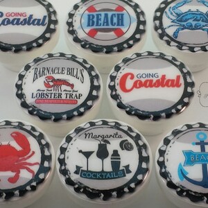 Peut inclure: Ensemble de boutons de placard en c&eacute;ramique blanche avec des motifs sur le th&egrave;me c&ocirc;tier. Les boutons pr&eacute;sentent des images de crabes, d'ancres, de cocktails et les phrases "Going Coastal" et "Beach". Chaque bouton a un design de bouchon de bouteille noir et blanc.