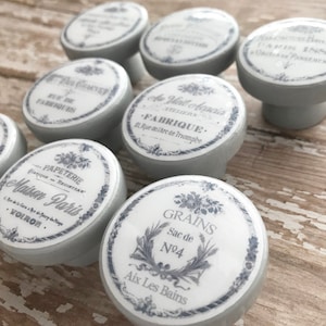 May include: A set of 10 decorative knobs with a light blue background and white lettering. The knobs feature French-inspired designs with text such as "Maison Paris", "Papeterie", "Grains", and "Aux Les Bains".