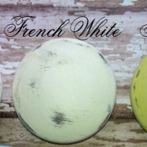 May include: Three paint samples on a wooden surface. The samples are labeled "Beach White", "French White", and "Tea Green".