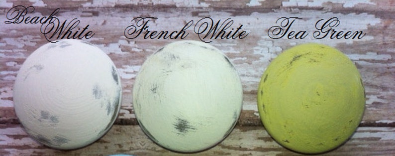 May include: Three paint samples on a wooden surface. The samples are labeled "Beach White", "French White", and "Tea Green".
