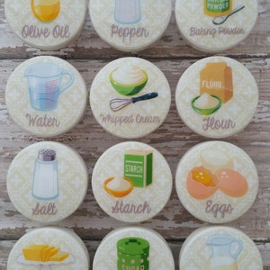 Könnte beinhalten: Zwölf runde, weiße Keramikknöpfe mit einem gelben und weißen Blumenmuster. Jeder Knopf zeigt eine andere Küchenzutaten-Illustration und eine Textbezeichnung: Olivenöl, Pfeffer, Backpulver, Wasser, Schlagsahne, Mehl, Salz, Stärke, Eier, Butter, Zucker und Milch.