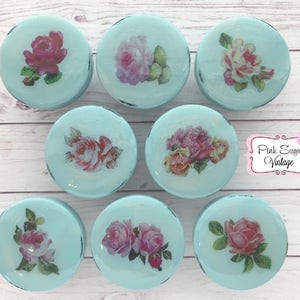 May include: Eight round, light blue, decorative boxes with a distressed finish. Each box features a different vintage floral design with pink, white, and green roses.