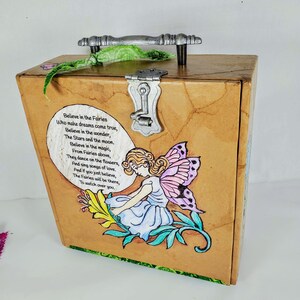 May include: A wooden box with a metal handle and a lock. The box is decorated with a fairy illustration and a poem about fairies. The poem reads: "Believe in the Fairies Who make dreams come true, Believe in the wonder, The Stars and the moon. Believe in the magic, From Fairies above, They dance on the flowers, And sing songs of love. And if you just believe, The Fairies will be there, To watch over you."