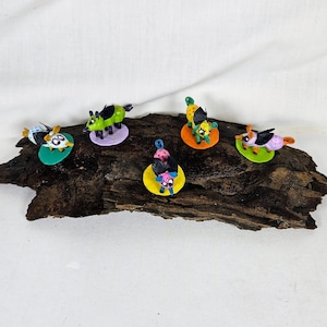 May include: Five colorful hand-painted ceramic animal figurines, each with a different design and color scheme. The figurines are standing on small round bases and are arranged on a piece of driftwood.