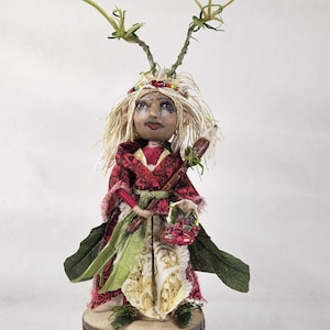 May include: A handcrafted doll with a whimsical design. The doll features a red and green patterned dress, blonde hair, and a decorative headpiece. It stands on a wooden base and holds a small basket of flowers and a staff.