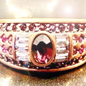 May include: A gold-toned ring with an oval, deep red gemstone at the center, flanked by rectangular and round clear and red stones. The band is encrusted with smaller red gemstones, creating a luxurious appearance.