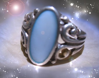 May include: A silver ring with an oval, light blue stone. The ring has an ornate, swirling design around the stone. The ring is set against a soft, white background with sparkling accents.