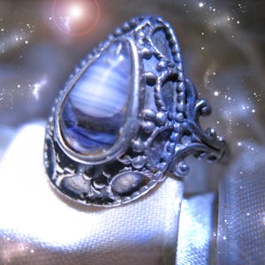 Op de afbeelding: Een sierlijke zilveren ring met een grote, druppelvormige steen met blauwe en witte strepen. De ring heeft een gedetailleerd, filigraan ontwerp rond de steen en langs de band, wat een vintage of antieke uitstraling creëert. De ring staat tegen een zachte, lichte achtergrond.