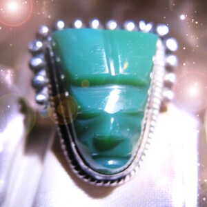 May include: A ring featuring a carved green stone shaped like a face, set in a silver bezel with a decorative beaded and twisted wire border. The stone has a smooth, polished surface with subtle facial features including eyes and a mouth.