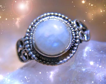 May include: A silver ring with a large, round, white moonstone set in a decorative bezel. The ring is set against a sparkly, out-of-focus background.