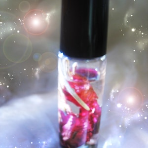 May include: A clear glass roll-on bottle with a black cap, filled with a liquid and red and pink flower petals. The background has a soft, blurred effect with white and light blue accents.