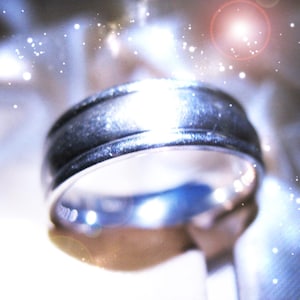 Op de afbeelding: Een zilverkleurige metalen ring met een gladde, gepolijste afwerking en een subtiele horizontale groef die in het midden van de band loopt. De ring heeft een afgerond profiel en reflecteert het licht, wat zorgt voor heldere accenten.