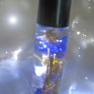 May include: A clear glass roller bottle with a black cap filled with a blue and white floral arrangement. The bottle is sitting on a white surface with a sparkly background.