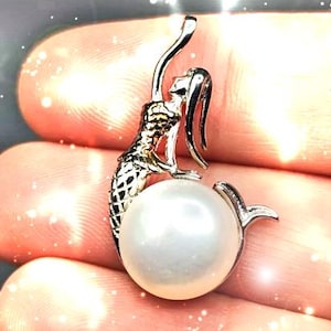 Puede incluir: Colgante de sirena plateada con una perla blanca grande y redonda. La sirena tiene escamas detalladas y cabello suelto, como si emergiera de la perla. El colgante está diseñado para ser usado como collar.