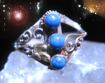 May include: A silver ring with three bright blue, round gemstones. The ring has an ornate design with floral and scroll details. The gemstones are set in a decorative setting.