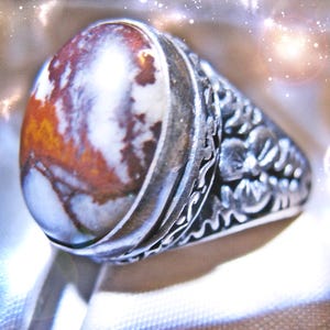 May include: A silver ring featuring a large, oval-shaped cabochon stone with a marbled pattern of white, brown, and rust-orange colors. The metal band is decorated with intricate, raised floral and leaf-like carvings along the sides.