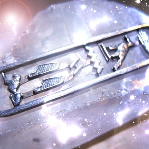 以下が含まれることがあります： エジプトのヒエログリフのデザインが施されたシルバーカラーのペンダント。ペンダントは、細部までこだわった人物像やシンボルが、細長い楕円形のフレームの中に配置されています。背景は、明るい色の、おそらく結晶質の表面です。