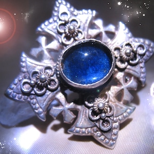Peut inclure: Broche argentée ornée d'une pierre centrale ovale bleu foncé. La broche présente un motif en étoile avec des détails floraux et en forme de croix. Le design est orné et détaillé, d'une esthétique vintage ou antique.