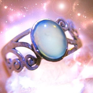 Op de afbeelding: Een zilverkleurige ring met een ovale, lichtblauwe edelsteen. De ring heeft een sierlijk, open ontwerp met wervelende details op de band. De edelsteen is gezet in een bezel setting en de achtergrond heeft een zacht, wazig effect.
