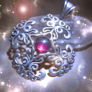 May include: A silver pendant with an intricate, swirling design. It features a central, round, deep pink gemstone. The metalwork has a detailed, open filigree pattern. The pendant is likely for a necklace.