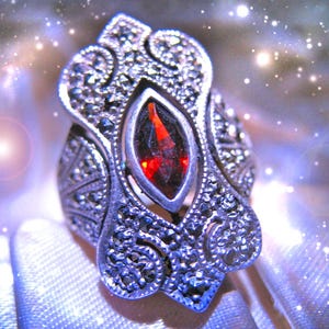 May include: A silver-toned ring featuring a central marquise-cut deep red gemstone surrounded by intricate metalwork and small dark grey stones. The metal surface displays a textured, ornate pattern with swirling details and a polished finish.