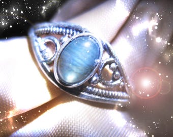 May include: A silver ring with an oval, light blue gemstone. The ring has an ornate, filigree design around the stone and along the band. The background is a soft, light-colored fabric, with sparkling effects.