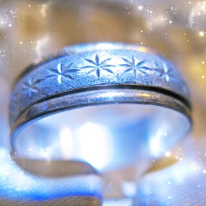 Op de afbeelding: Een zilverkleurige metalen ring met een centrale band voorzien van een herhalend gegraveerd sterpatroon. De ring heeft een gladde, gepolijste afwerking langs de randen en een getextureerd, mat uiterlijk in het midden waar de sterren zijn gegraveerd.