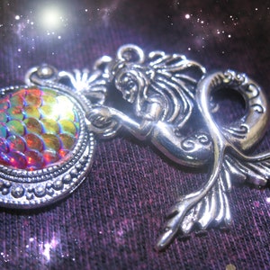 May include: A silver-toned mermaid pendant with a round, iridescent, scale-like center. The mermaid is holding the round element. The pendant is set against a dark, textured background with a starry effect, creating a fantasy theme.