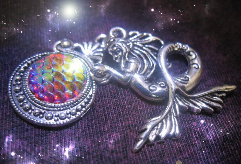 May include: A silver-toned mermaid pendant with a round, iridescent, scale-like center. The mermaid is holding the round element. The pendant is set against a dark, textured background with a starry effect, creating a fantasy theme.