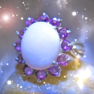 Puede incluir: Un anillo de plata con una gran piedra de ópalo blanco rodeada por un halo de pequeñas piedras preciosas púrpuras.