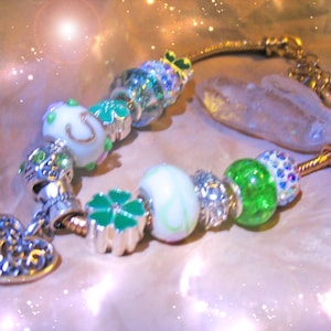 May include: A gold-toned bracelet adorned with various charms. The charms include a heart-shaped pendant, a four-leaf clover, and beads in shades of green, white, and silver. Some beads are encrusted with sparkling crystals. A clear, oblong stone is also present.