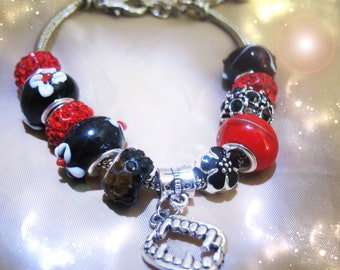 May include: Silver charm bracelet with a variety of beads in black, red, and silver. The beads include red crystal-covered spheres, black beads with white floral designs, and a red, glossy bead. A silver charm of vampire fangs hangs from the bracelet.