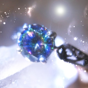 May include: A close-up of a ring with a large, iridescent gemstone. The stone displays a spectrum of colors, including blues, greens, and purples. The ring's band is a dark metal, and the setting has small prongs holding the stone.